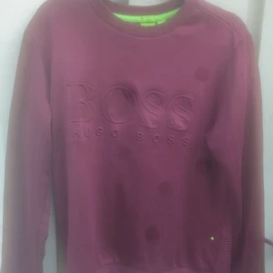 Vinröd sweatshirt från Hugo Boss - Säljer en vinröd sweatshirt från Hugo Boss med stort BOSS-tryck i relief på bröstet. Tröjan har rund hals, långa ärmar och ribbade muddar. Baksidan är ribbad för en extra snygg detalj. Perfekt för dig som gillar stilrena märkesplagg.