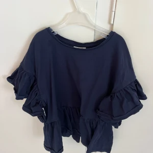 Mörkblå volangblus med kort ärm - Supersöt mörkblå blus med volanger både på ärmarna och nertill. Den har en lös passform och är tillverkad i mjuk bomull. Rund halsringning och korta ärmar med extra volym för en cool look.