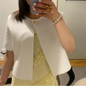 Vit kort kavaj med knapp - Snygg vit kavaj med kort ärm och rak passform. Jackan har en stilren design med en dekorativ guldig knapp upptill och öppen framtill. Perfekt för att lyfta en outfit med en clean och fräsch look.