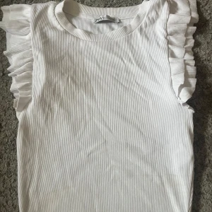 Vit ribbad topp med volangärmar Zara - Supersöt vit ribbad topp från Zara med volangdetaljer på axlarna. Ärmlös modell som ger en fräsch och trendig look. Perfekt att styla med jeans eller kjol för en clean vibe.