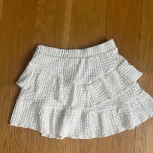 Vit volangkjol från Zara - Supersöt vit kortkjol från Zara med flera volanger och smockad struktur. Kjolen har en luftig känsla och är perfekt för varma dagar. Elastisk midja för extra komfort och snygg passform.💕Passar även xs och xxs.