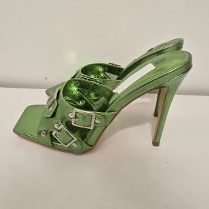 Gröna metallic klackpumps med spännen - Statement-pumps i metallicgrönt med flera silvriga spännen och nitar. Skorna har fyrkantig tå, hög klack och öppen häl. Perfekta för dig som vill sticka ut med en edgy och trendig look.