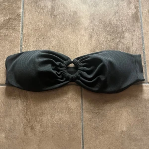 Svart bandeau bikiniöverdel - Snygg svart bandeau bikiniöverdel med ribbad struktur och en cool ringdetalj i mitten som ger en trendig look. Perfekt för stranden eller poolen, enkel att matcha med olika underdelar. Stilren och modern design. Storleken är S/M, passar någon som har mellan B/C kupa