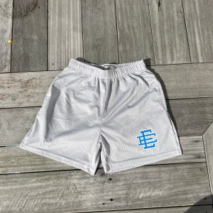 Gråa meshshorts från Eric Emanuel - Snygga grå meshshorts från Eric Emanuel med blå logga. Shortsen har elastisk midja och är tillverkade i luftig mesh, perfekta för varma dagar eller sport. Klassisk loose fit och sportig vibe.   Storlek: M men passar som S. Längd: 43cm