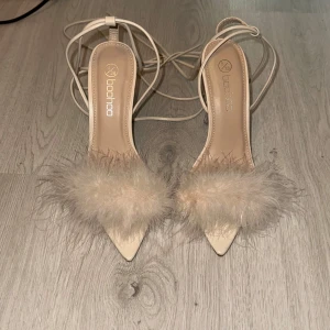 Beiga Boohoo klackar med fjädrar - Snygga beiga klackar från Boohoo med spetsig tå, transparent hög klack och fluffiga fjädrar framtill. Skorna har tunna remmar som knyts runt vristen för en trendig look. 