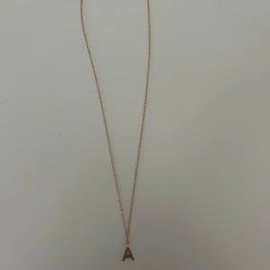 Guldigt halsband med bokstav A - Stilrent halsband i guld med en tunn kedja och ett hänge i form av bokstaven A. Perfekt accessoar för dig som vill ha något personligt och enkelt men ändå snyggt. Passar till alla outfits och är lätt att matcha med andra smycken.