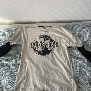 Beige t-shirt med svart tryck - Säljer en beige t-shirt med svart grafiskt tryck och texten 'individualex' på bröstet. T-shirten har svarta långa ärmar under de korta, vilket ger en lager-på-lager-look. Materialet känns som bomull och passformen är normal.