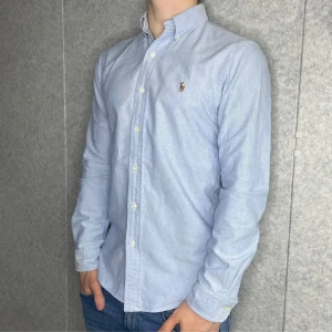 Ljusblå skjorta från Polo Ralph Lauren - Säljer en stilren ljusblå skjorta från Polo Ralph Lauren i slim fit. Skjortan har klassisk button-down krage, broderad logga på bröstet och vita knappar. Tillverkad i mjuk bomull och passar perfekt till jeans eller chinos.