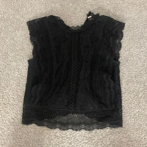 Svart spetsblus med volangdetaljer - Supersnygg svart blus i spets med breda volangdetaljer längs ärmhålen och nederkanten. Blusen är ärmlös och har ett romantiskt mönster i spetsen som ger en cool och elegant vibe. Perfekt att styla med jeans eller kjol för en trendig look.