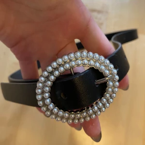 Svart skärp med pärlspänne - Snyggt svart bälte med ett ovalt spänne dekorerat med två rader pärlor i silverfärg. Bältet är stilrent och har en klassisk form som passar till jeans eller klänning. Materialet är slätt och ger en cool vibe till din outfit.