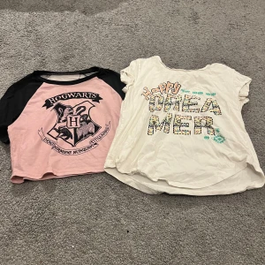 2 t-shirts: Hogwarts & Happy Dreamer - Två t-shirts säljes! En rosa och svart Hogwarts t-shirt med coolt tryck och korta ärmar, croppad passform. Den andra är vit med färgglatt 'Happy Dreamer'-tryck och mönstrade bokstäver, också kortärmad och loose fit. Perfekt för dig som gillar unika prints och chill stil.