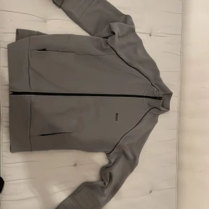 Grå vindjacka från BOSS. Originellt pris 1,200 - Snygg grå tröja från BOSS med stilren design och svarta detaljer. Jackan har hel dragkedja framtill, diskret logga på bröstet och praktiska fickor med dragkedja. Materialet känns lätt och smidigt, perfekt för dig som gillar minimalistisk stil. Bara används runt 6 gånger, andledningen helt enkelt för jag säljer, inte min stil längre! Ps kan nog också passa XL