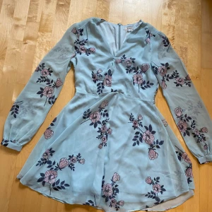 Blommig ljusblå klänning Glamorous - Supersöt ljusblå kortklänning från Glamorous med rosa och mörkblå blommor. Klänningen har långärmade transparenta ärmar, v-ringad hals och dragkedja i ryggen. Materialet är lätt och flowy, perfekt för vår och sommar. Passformen är normal och kjolen är lätt utsvängd.