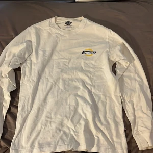 Vit långärmad Dickies tröja - Vit långärmad tröja från Dickies i bomull med klassisk rund hals. Stor Dickies-logga i gult och svart på ryggen och mindre logga på bröstet. Perfekt basic med streetkänsla och relaxed fit.