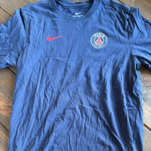 Blå PSG t-shirt från Nike - Snygg blå t-shirt från Nike med Paris Saint-Germain-logga på bröstet och röd Nike swoosh. Klassisk rund hals och korta ärmar. Perfekt för dig som gillar fotboll och vill visa vilket lag du hejar på.