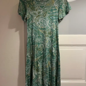 Grön paisleymönstrad klänning  - Snygg grön kortklänning med paisleyinspirerat mönster i grönt, vitt och beige. Klänningen har rund hals, korta ärmar och ett mjukt, flowy fall. Perfekt för dig som gillar boho-vibe och färg. Materialet är lätt och känns skönt mot huden.