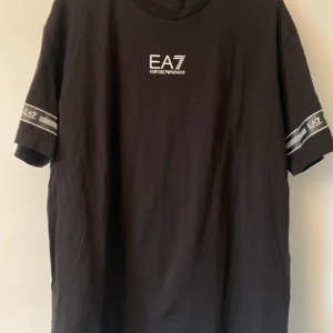Svart EA7 Emporio Armani t-shirt - Svart t-shirt från EA7 Emporio Armani med vit logga på bröstet och tryck på ärmarna samt nedtill. Klassisk rund hals och rak passform. Tillverkad i 100% bomull, perfekt för dig som gillar stilrena och snygga märkesplagg.
