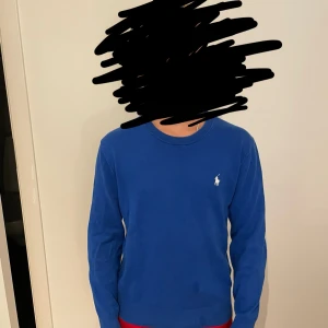 Blå tröja från Polo Ralph Lauren - Snygg blå tröja från Polo Ralph Lauren med klassisk rund hals och vit broderad logga på bröstet. Tröjan har långa ärmar och ribbade muddar vid ärmslut och nederkant. Perfekt för dig som gillar stilrena och tidlösa plagg. Den har tyvärr ett litet hål på höger armbåge men det syns inte när man har på sig den och det är nog enkelt att laga. Köpt på NK för 800!