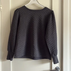 Mörkgrå quiltad tröja Jacqueline de Yong - Quiltad mörkgrå tröja från Jacqueline de Yong i storlek L. Tröjan har långärmade puffärmar, breda muddar och rund halsringning. Perfekt för dig som gillar en stilren och chill look med lite struktur i tyget.