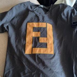 Svart Fendi t-shirt med stor logga - Svart t-shirt från Fendi med en stor, kontrasterande Fendi-logga i brunt på bröstet. Klassisk rund hals och korta ärmar. Tillverkad i 100% bomull för skön och mjuk känsla. Perfekt för dig som gillar exklusiva streetwear-vibes och tydliga designerloggor. Aldrig använd 