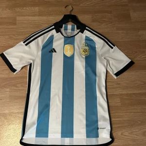 Argentina VM 2022 fotbollströja Adidas - Säljer en officiell Argentina VM 2022 fotbollströja från Adidas i storlek 15-16 år/176. Tröjan har klassiska blå och vita vertikala ränder, svarta detaljer vid krage och ärmslut samt svarta Adidas-ränder på axlarna. Broderade FIFA World Champions 2022 och AFA-emblem på bröstet. Tillverkad i lätt polyester. Säljer då jag växt ur den.