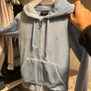 Ljusblå Juicy Couture set - Säljer en ljusblå hoodie och byxor från Juicy Couture med dragkedja och huva. Tröjan har långa ärmar, snörning i huvan och en stor ficka framtill. Materialet är mjuk polyester med lite stretch för extra komfort. Perfekt för chill dagar eller när du vill ha en snygg streetstyle-look.