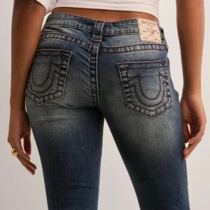 True Religion bootcut jeans blå - Säljer ett par blå low waist jeans från True Religion med bootcut passform. Jeansen har röda kontrastsömmar. Dom är sydda vid midjan men det går att sprätta bort.  Skriv om ni vill att jag ska ta bort det jag har sytt runt midjan!!! Storlek 28 vilket ungefär motsvaras storlek S/36. Nya pris är 1 299
