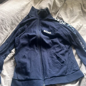 Mörkblå zipjacka från Boss - Mörkblå zipjacka från Boss med hög krage och tryckt logga på bröstet och ärmen. Jackan har två fickor framtill och är tillverkad i mjuk bomull. Perfekt för en chill och sportig look.