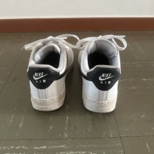 Nike Air Force 1 vita och svarta sneakers - Nike Air Force 1 sneakers i klassisk vit färg med svarta detaljer och swoosh. Skorna har snörning, perforerad tå och platt sula. Tillverkade i skinn med textilfoder för extra komfort. Perfekt för dig som gillar streetstyle och tidlös design.