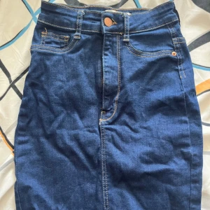 Blå jeanskjol från Gina Tricot XS - Säljer en klassisk blå jeanskjol från Gina Tricot, modell Molly. Kjolen har normal passform, synliga fickor fram och bälteshällor. Snygga kontrastsömmar och knappstängning framtill. Perfekt för dig som gillar enkel och tidlös denimstil.