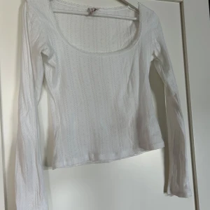 Vit långärmad topp från Nelly - Säljer en vit långärmad topp från Nelly i storlek XS. Toppen har en djup, fyrkantig urringning och är gjord i ett mjukt, stretchigt material med diskret hålmönster och spetskant🥰