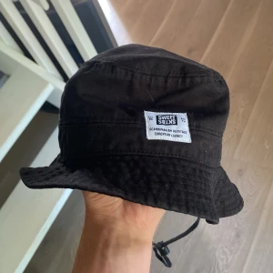 Svart bucket hat från Sweet SKTBS - Cool svart bucket hat från Sweet SKTBS med vit patch framtill där det står 'Scandinavian Heritage European Lunacy'. Hatten har klassisk form med nedvikt brätte och justerbar hakrem. Perfekt accessoar för en avslappnad streetstyle.