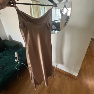 Champagnefärgad satinklänning med smala band - Säljer en elegant långklänning i champagnefärgad satin med smala axelband och snygg draperad urringning. Klänningen har en slät siluett och faller mjukt längs kroppen, perfekt för dig som gillar minimalistisk och stilren look.