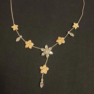 Blommigt halsband i guld och silver - Supersött halsband med små blommor i guldton och glittriga silverdetaljer. Blommorna har en kristall i mitten och halsbandet har en elegant Y-formad design med extra hängande blomma och kristall. Perfekt för att ge din outfit en romantisk touch.
