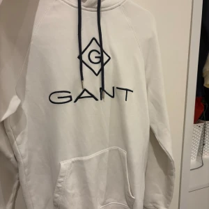 Vit hoodie från GANT med logga - Vit hoodie från GANT i storlek XS med broderad marinblå logga på bröstet. Tröjan har huva med snörning, känguruficka och långa ärmar. Mjuk bomullskvalitet som är skön att ha på sig. Perfekt för chill dagar eller när du vill ha en clean look.