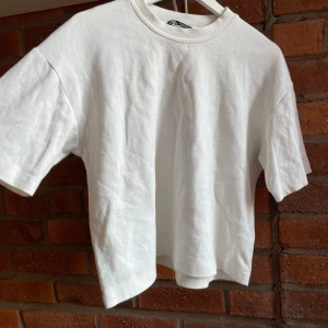 Vit croppad t-shirt från Zara - En clean vit t-shirt från Zara i croppad modell med rund hals och korta ärmar. Tillverkad i mjuk bomull som känns skön mot huden. Perfekt för dig som gillar enkel och stilren vibe. Passar dig som vill ha en lite loose fit.