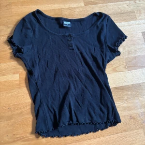 Svart ribbad topp Urban Classics S - Svart ribbad topp från Urban Classics i storlek S. Modellen har korta ärmar med söt volangkant, rund halsringning och tre knappar framtill. Materialet är mjukt och stretchigt, perfekt för en chill och stilren look.
