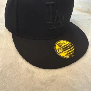 Svart LA keps New Era HELT NY - Svart New Era 59FIFTY keps med LA-logga broderad framtill i samma svarta tön. Klassisk flat skärm och guldfärgad sticker på skärmen. Helt ny aldrig använd. Kommer med pappret inuti kepsen också.