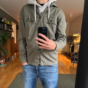 Olivgrön overshirt från 157, stl S - Säljer en olivgrön overshirt från 157 med klassisk krage och två stora bröstfickor med knappar. Jackan är i bomull och har en clean, avslappnad look som passar perfekt till jeans. Långärmad och stängs med knappar framtill.