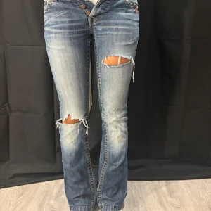  bootcut jeans med slitningar - Snygga lågmidjade jeans i ljusblå tvätt med slitna detaljer och hål på båda knäna. Bootcut-modell med klassisk femficksdesign och knappgylf. Jeansen har en cool, avslappnad vibe och är tillverkade i äkta denim. Storlek 24