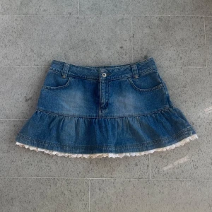 Jeanskjol med volang och spetskant - Säljer en blå jeanskjol med volang och vit spetskant längst ner. Kjolen har klassiska fickor fram och bak, bälteshällor och knappstängning. Den har en kort och utsvängd modell som ger en söt och trendig vibe. Midjemått: 38cm. Använd gärna köp nu❤️