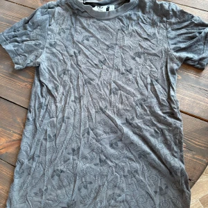 Grå Adidas t-shirt med logomönster - Säljer en grå t-shirt från Adidas med all-over tryck av Adidas logga och text. T-shirten har korta ärmar och rund hals. Perfekt för dig som gillar sportig och street-inspirerad stil. Materialet är mjuk bomull som känns skönt mot huden.