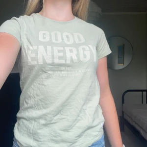 Ljusgrön t-shirt med tryck från 157 - Ljusgrön t-shirt från 157 med stort vitt tryck på bröstet där det står 'GOOD ENERGY'. Klassisk rund halsringning och korta ärmar. Skön och mjuk i bomull, perfekt till jeans eller shorts. Enkel och clean stil med en positiv vibe.