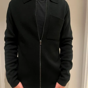 Svart overshirt med dragkedja från Jack n Jones  - Stilren svart overshirt med lång ärm och rak passform. Jackan har en stor bröstficka och hel dragkedja framtill. Perfekt för lager-på-lager och funkar till många olika outfits. Materialet är mjukt och känns skönt mot huden. Jätte bra skick aldrig använd. 