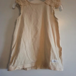 Ljusgul ärmlös topp med spets från Newbie - Supersöt ljusgul topp från Newbie i mjuk bomull. Ärmlös modell med broderade spetsdetaljer på axlarna och en liten knapp i nacken. Perfekt för varma dagar och enkel att matcha med allt. Storlek 98/104.