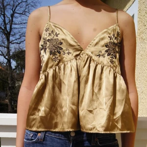 Guldig blommig linnetopp från Zara - Supersöt guldig linnetopp från Zara med tunna axelband och djup v-ringning. Toppen har broderade svarta blommor framtill och är lätt utsvängd nertill för en luftig känsla. Perfekt för varma dagar när du vill sticka ut lite extra.