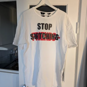 Vit VLONE t-shirt med tryck, strl L - Vit t-shirt från VLONE i bomull med stora tryck fram och bak. Framsidan har texten 'STOP SNITCHING' i svart och rött, baksidan har en stor röd V med 'NO CAP' i svart. Klassisk passform och rund hals. Perfekt för dig som gillar streetwear och statement-plagg.