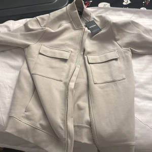 Beige bomberjacka från Divided - Snygg beige bomberjacka från Divided med två stora fickor framtill och dragkedja hela vägen. Jackan har ribbade muddar vid ärmslut och midja, samt en stilren, minimalistisk look. Perfekt för dig som gillar clean och modern stil.