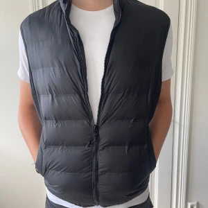 Svart dunväst från Zara - Säljer en svart dunväst från Zara i storlek M. Västen har en stilren look med quiltad design, hög krage och dragkedja framtill. Perfekt att slänga över en hoodie eller t-shirt för extra värme. Enkel och clean stil som funkar till det mesta.