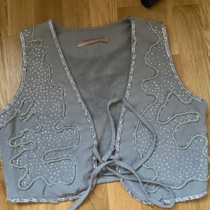 Grå väst med paljetter rave bohem hippie - Snygg grå väst från Juicy Couture med paljett- och pärldetaljer samt dekorativa broderier. Västen har knytband framtill och är fodrad med mjuk fleece på insidan. Perfekt för att ge outfiten en unik touch.